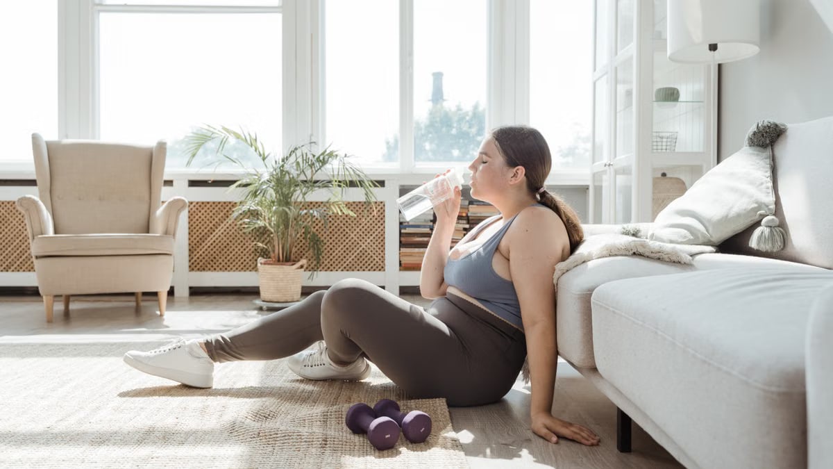 Curvy woman in activewear, drinking water in a sunny living room with dumbbells nearby.